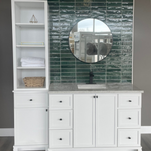 Modern white bathroom vanity with a storage tower, dark green subway tile backsplash, and a circular black-framed mirror.